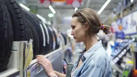 Department-of-car-accessories-in-the-store.-Large-hypermarket.-The-girl-is-standing-near-the-rack-with-the-tires.-Selects.-Blurred-store-background