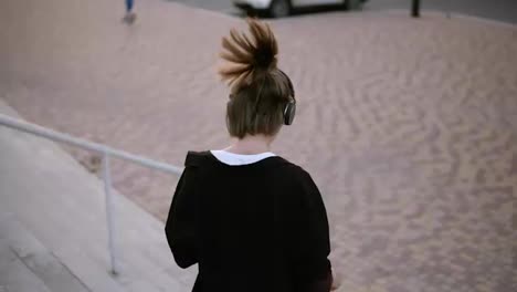Happy-stylish-woman-with-ponytail-going-down-the-stairs-playfully-in-the-park.-Whirling-and-dancing-with-music-in-her-headphones