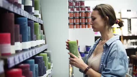 Side-view-of-an-attractive-woman-choosing-different-colorful-candles-from-the-rack-in-supermarket.-Smelling-green-candle-from-the-shelf