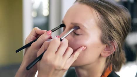 The-professional-make-up-artist-does-make-up-for-the-young-cute-model.-Artist-putting-and-eyes-shadows-using-two-brushes-and-does
