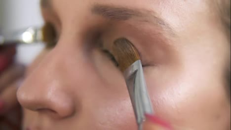 Make-up-artist-applying-light-brown-shadows-on-and-eyelids-with-two-brushes-together,-Close-up-of-a-young-caucasian-woman's-eyes