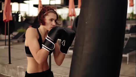 Young-athletic-female-boxer-in-gloves-kicking-a-boxing-bag-with-her-hands-and-elbows.-Workout-outside.-Female-boxer-training-in-boxing-gloves.-Shot-in-4k