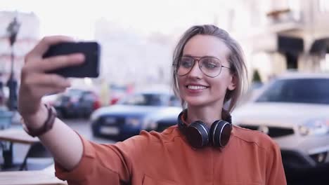 Cheerful-attractive-female-dressed-in-orange-shirt-and-sunglasses-making-selfie-on-smartphone-camera,-posing,-standing-on-street
