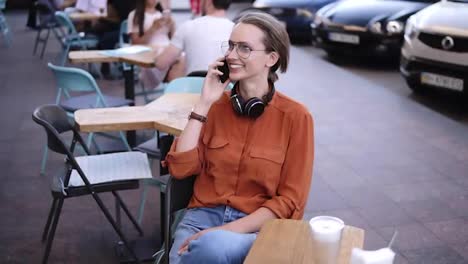 Happy,-confident-blonde-woman-in-denim-and-orange-shirt-sitting-on-a-chair-in-a-street-cafe.-Talking-by-her-smartphone.-Smiling