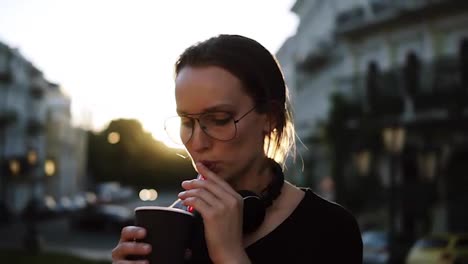 Young-woman-in-stylish-sunglasses-drinking-a-coffee-with-plastic-straws-on-the-street.-Evening-dusk.-son-shines-on-the