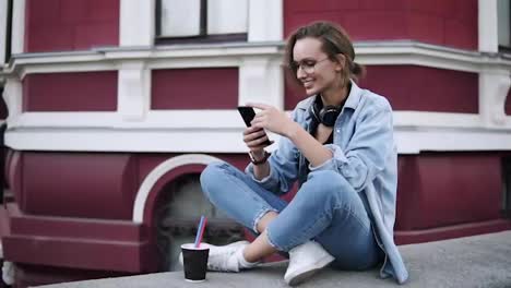 A-fashionable-girl-in-glasses-sits-on-the-street-on-a-parapet,-concentrates-on-her-mobile-phone.-Smiling,-communication