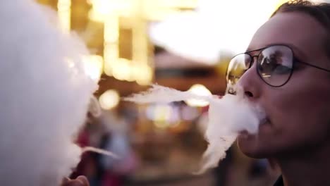 Beautiful-girl-taking-cotton-candy-in-and-amusement-park-near-the-blurred-carousel.-Wearing-trendy-glasses,-happy,-smiling.-Close