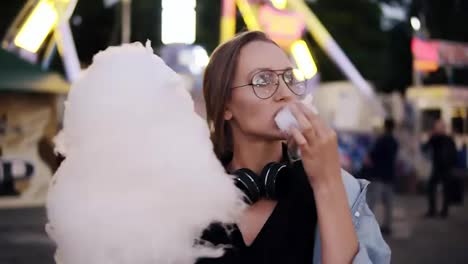 and-attractive-girl-walking-in-the-amusement-park,-eats-white-cotton-candy.-Girl-wearing-casual-clothes,-headphones-on-her-neck