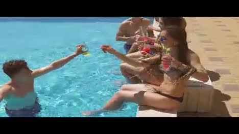 Happy-young-friends-hanging-out-with-coctails-and-chatting-at-the-side-of-the-pool-in-the-summertime,-drinking-cocktails-and-doing-cheers.-Pool-party.-Shot-in-4k