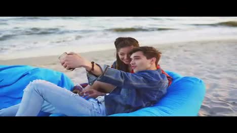 An-couple-having-fun-and-taking-selfies-laying-on-easy-chairs-with-blanket-on-the-beach.-Shot-in-4k.