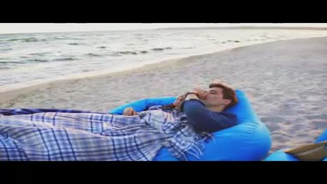 Couple-laying-on-easychairs-on-the-beach-covered-with-blanket.-Man-smoking-electronic-cigarette.-Vaper.-Shot-in-4k