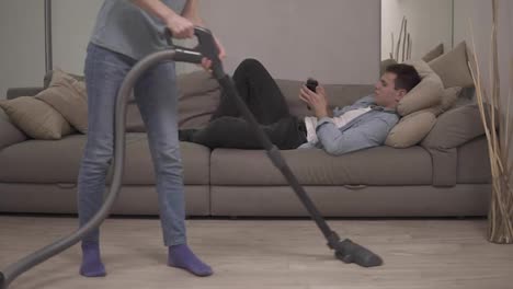 Unrecognizable-woman-in-jeans-vacuuming-parquet-floor,-and-her-husband-plays-on-smartphone-on-couch.-Front-view,-slow-motion