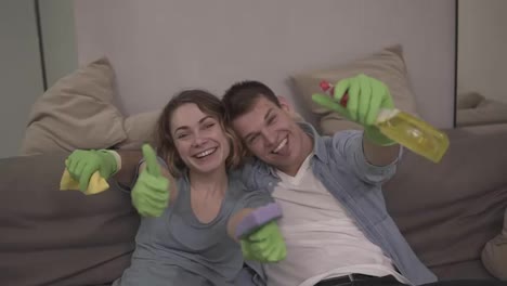 Portrait-of-a-young,-cheerful-couple-in-green-rubber-gloves.-The-family-finishing-the-house-cleaning-together.-Happy-tired-team,-sitting-on-a-couch,-looking-to-the-camera,-smiling,-thumbs-up