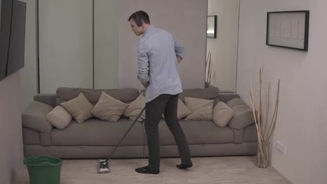 People,-housework-and-housekeeping-concept---happy-man-in-headphones-with-mop-cleaning-floor-and-dancing-at-home.-Grey-couch-on-the-background