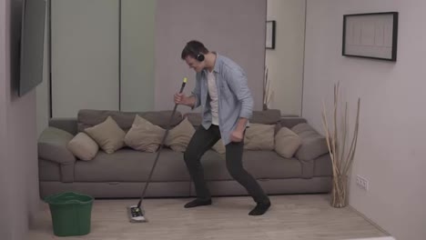 People,-housework-and-housekeeping-concept---happy-man-in-headphones-with-mop-cleaning-floor-and-dancing-at-home.-Using-swab-pipe-as-a-guitar.-Grey-couch-on-the-background