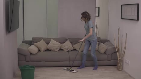 People,-housework-and-housekeeping-concept---happy-woman-in-headphones-with-mop-cleaning-floor-and-dancing-at-home.-Grey-couch-on-the-background.-Slow-motion