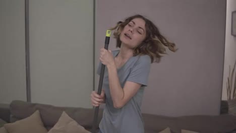 People,-housework-and-housekeeping-concept---happy-woman-in-headphones-with-mop-cleaning-floor-and-dancing-at-home.-Using-swab-pipe-as-a-microphone.-Grey-couch-on-the-background