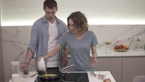 Cheerful-couplemaking-breakfast-together.-Girl-pouring-mixed-eggs-from-a-bowl-to-a-hot-pot-on-a-kitchen-stove.-Husband-standing-behind-and-watching.-Slow-motion