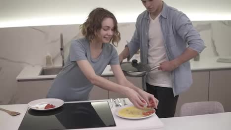 Cheerful-couple-on-bright,-white-kitchen.-They-prepared-omelette-on-pan,-girlfriend-is-serving-dish,-adding-tomatos.-Excited-and-hungry.-Slow-motion