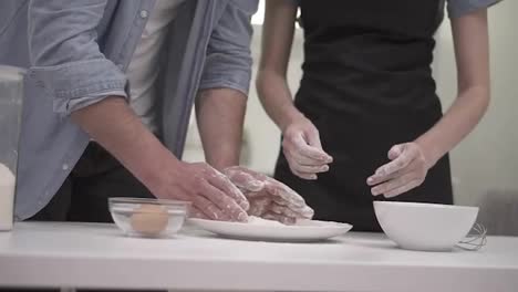 Attractive-caucasian-man-preparing-dough-on-the-white-counter-on-kitchen.-His-girlfriend-helping-him-with-that.-Young-couple-having-fun,-faces-in-flour.-Slowmotion-shot