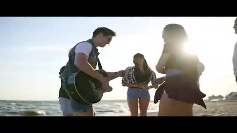 Summer-party-on-the-beach.-Young-friends-drinking-cocktails,-dancing-in-the-cirkle,-playing-guitar,-singing-songs-and-clapping-on-a-beach-at-the-water's-edge-during-the-sunset.-Slowmotion-shot