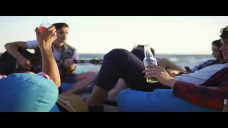 Group-of-friends-drinking-cocktails-and-beer-and-doing-cheers-sitting-on-easychairs-on-the-beach-and-listening-to-a-friend-playing-guitar-on-a-summer-evening-during-a-sunset.-Slowmotion-shot