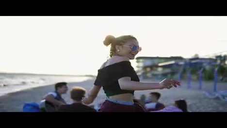 Carefree-young-hipster-woman-with-dreads-dancing-in-the-sunset-with-group-of-friends-sitting-on-easychairs-on-the-beach-and-playing-guitar-on-a-summer-evening-during-a-sunset.-Slowmotion-shot
