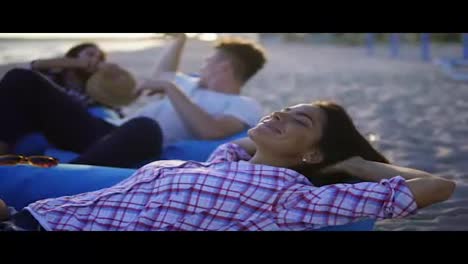 Happy-young-woman-laying-on-easy-chair-and-smiling-among-group-of-friends-on-the-beach-during-a-sunset.-Slowmotion-shot