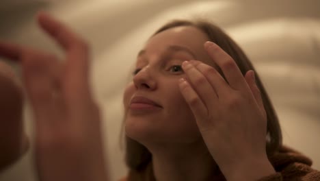 Young-beautiful-light-brown-haired-girl-is-applying-wrinkle-cream-on-eyelid-looking-in-the-mirror.-Handheld-footage