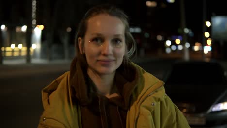 Gorgeous-front-footage-of-a-naturally-beautiful-young-woman-with-fair-hair-standing-on-the-street-and-smiling.-Night-time