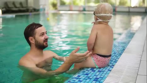 Young-father-is-showing-to-his-little-son-to-dive-under-water-in-the-swimming-pool-while-he-is-sitting-on-the-border.-He-is-jumping-in,-swimming,-then-his-mother-is-lifting-him-up-from-the-water