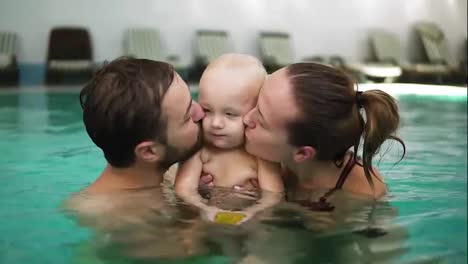 Young-mother-and-father-are-holding-their-cute-child-in-their-hands-in-the-swimming-pool-and-kissing-him-from-both-sides.-Happy-child-is-smiling