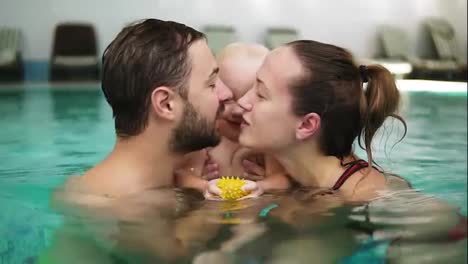 Young-mother-and-father-are-holding-their-cute-child-in-the-swimming-pool-and-kissing-each-other-in-their-lips.-Happy-child-is-smiling