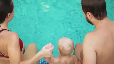 Back-view-of-young-mother-and-father-together-with-their-child-jumping-in-the-swimming-pool-holding-hands.-Mother-and-father-are-teaching-their-kid-how-to-swim