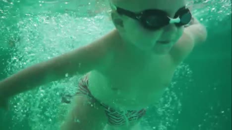 An-underwater-shot-of-a-cute-blonde-toddler-in-protective-glasses-swimming-under-the-water-in-the-swimming-pool.-Slowmotion