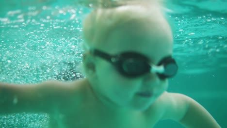 Und-Unterwasseraufnahme-Eines-Süßen-Blonden-Kleinkindes-Mit-Schutzbrille,-Das-Im-Schwimmbecken-Unter-Wasser-Schwimmt.-Zeitlupe
