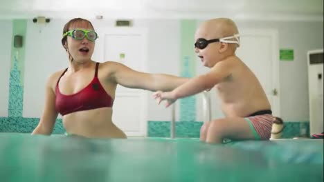 Cute-blonde-toddler-in-protective-glasses-is-diving-under-the-water-together-with-his-mother-in-the-swimming-pool-trying-to-take-out-his-toy.-His-mother-is-teaching-him-how-to-swim.-An-underwater-shot
