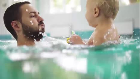 Young-father-is-lifting-his-little-boy-from-the-water-while-teaching-him-how-to-swim-in-the-swimming-pool.-Happy-little-boy-and-his-father-are-laughing-and-having-fun