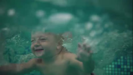 Cute-blonde-toddler-is-jumping-under-the-water-in-the-swimming-pool-and-swimming-there-until-his-mother-is-lifting-him-from-the-water.-An-underwater-shot