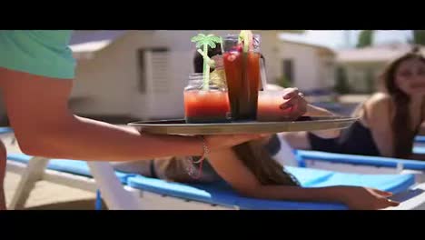 An-unrecognizable-waiter-bringing-cocktails-for-beautiful-young-girls-relaxing-by-the-pool.-Summertime-pool-party.-Slowmotion-shot