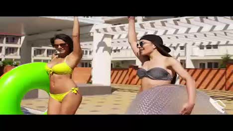 Two-attractive-girls-in-sunglasses-walking-wih-inflatable-tubes-by-the-pool-and-smiling.-Pretty-women-having-a-pool-party.-Slowmotion-shot