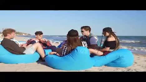 Group-of-friends-sitting-on-easychairs-on-the-beach-playing-guitar-and-singing-on-a-summer-evening.-Slowmotion-shot