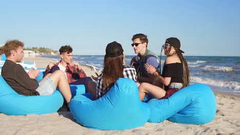 Group-of-friends-sitting-on-easy-chairs-on-the-beach-playing-guitar-and-singing-on-a-summer-evening.-Slow-Motion-shot