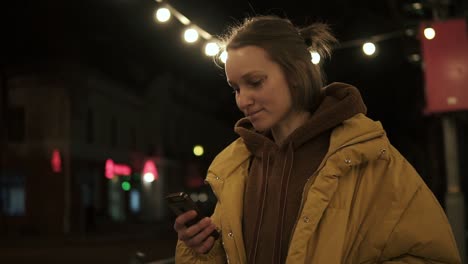 Pretty-girl-in-the-middle-of-a-dark-street-with-lights-looks-at-the-phone-and-smiles.-Yellow-coat.-Side-view