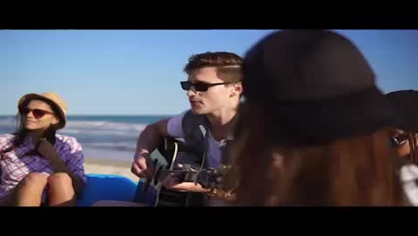 Young-man-playing-guitar-among-group-of-friends-sitting-on-easychairs-on-the-beach-and-singing-on-a-summer-evening.-Slowmotion-shot