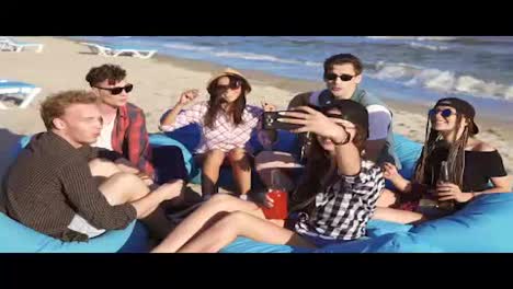Young-hipster-girl-recording-a-video-or-taking-selfie-of-group-of-friends-sitting-on-easychairs-on-the-beach,-playing-guitar-and-singing-on-a-summer-evening.-Slowmotion-shot