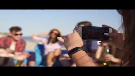 Closeup-view-of-a-young-hipster-girl-recording-a-video-or-taking-pictures-of-group-of-friends-sitting-on-easychairs-on-the-beach,-playing-guitar-and-singing-on-a-summer-evening.-Slowmotion-shot.