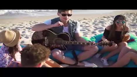 Junger-Mann-Spielt-Gitarre-In-Einer-Gruppe-Von-Freunden,-Sitzt-Auf-Sesseln-Am-Strand-Und-Singt-An-Einem-Sommerabend.-Zeitlupenaufnahme