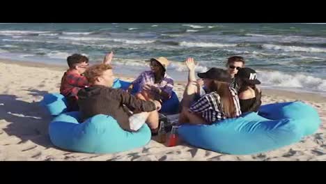 Eine-Gruppe-Junger-Freunde-Sitzt-Am-Strand,-Hat-Spaß-Und-Gibt-Sich-Gegenseitig-High-Fives-An-Einem-Sommerabend-In-Zeitlupe