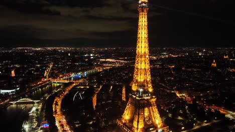 Eiffel-Tower-summit-with-automated-light-beams-sparkling,-Paris-cityscape-and-skylines-at-night,-Aerial-view